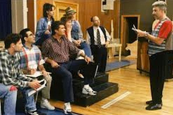 photo of a scene from the film "Waiting For Guffman" featuring the cast & director of a fictitious musical.